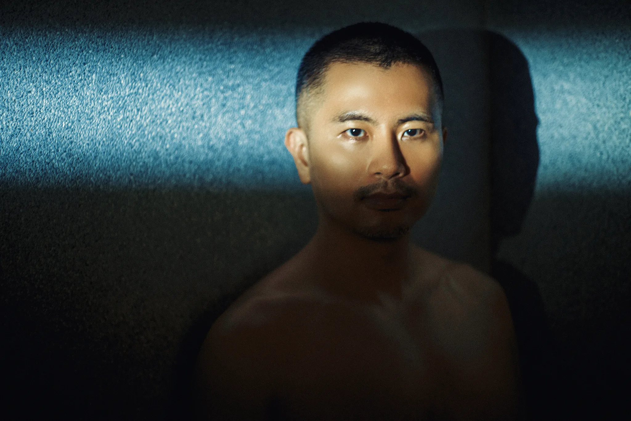 男子正面凝視鏡頭，背景呈現神秘的藍色光帶與強烈陰影，為宇宙拿鐵極具故事感的男體攝影作品。